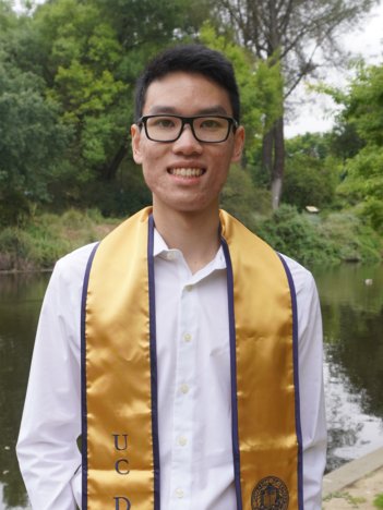 A young asian man with short hair and black-rimmed glasses wears a light pink button-down and a gold UC Davis graduation stole and smiles in front of a lush green background.